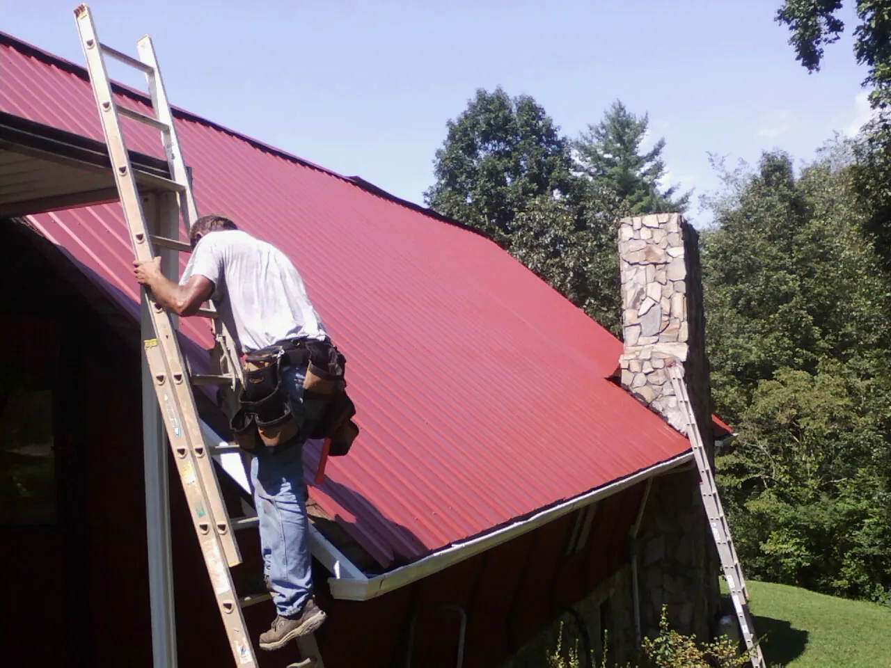 Roofer on a ladder beside a red metal roof and stone chimney — Soffit & Fascia Repair by Roofing Brothers in Bear Mountain